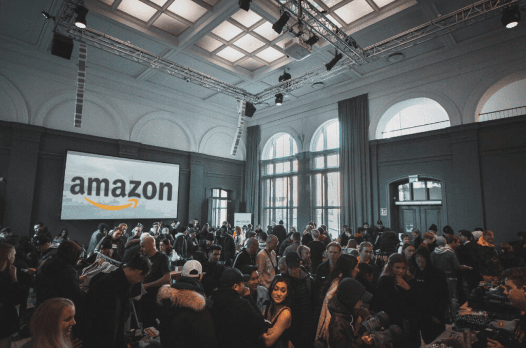 Crowded indoor event with a large Amazon logo on screen.