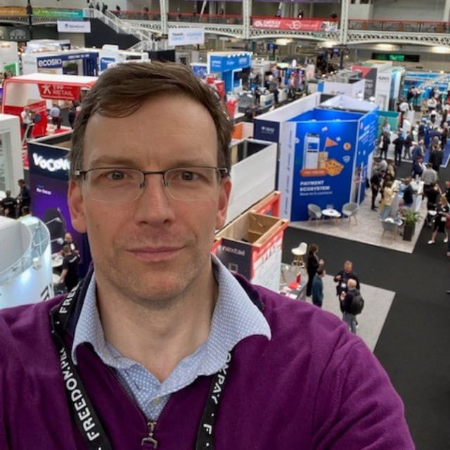 Man in a purple sweater at a busy trade show.