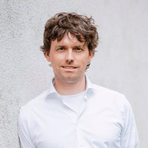 A man with messy brown hair wearing a white shirt.