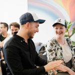 Two coworkers in trucker hats laughing during an office meeting.