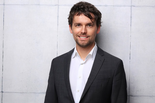 Smiling man in a suit jacket against a concrete wall