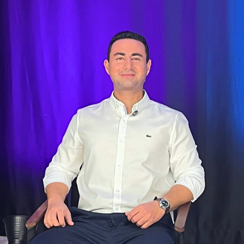 Man in white shirt sitting before purple backdrop.