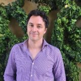 Man in purple checkered shirt against a leafy green wall.