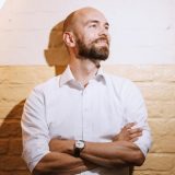 Bearded man in white shirt against a brick wall.